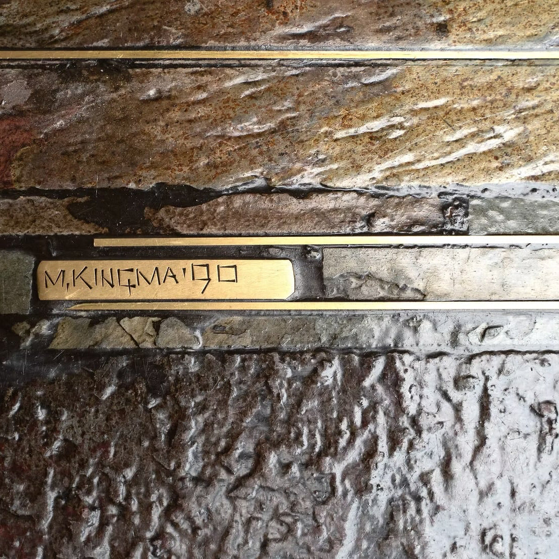 A close-up of a rectangular brass plaque engraved with 'M. KINEMA 1990' in black lettering, mounted on a textured stone wall.