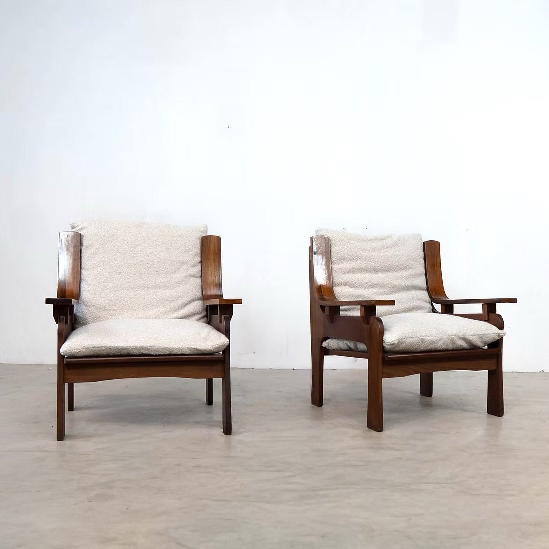 Two mid-century modern wooden armchairs with cream-colored cushions, set against a minimalist white wall and concrete floor.