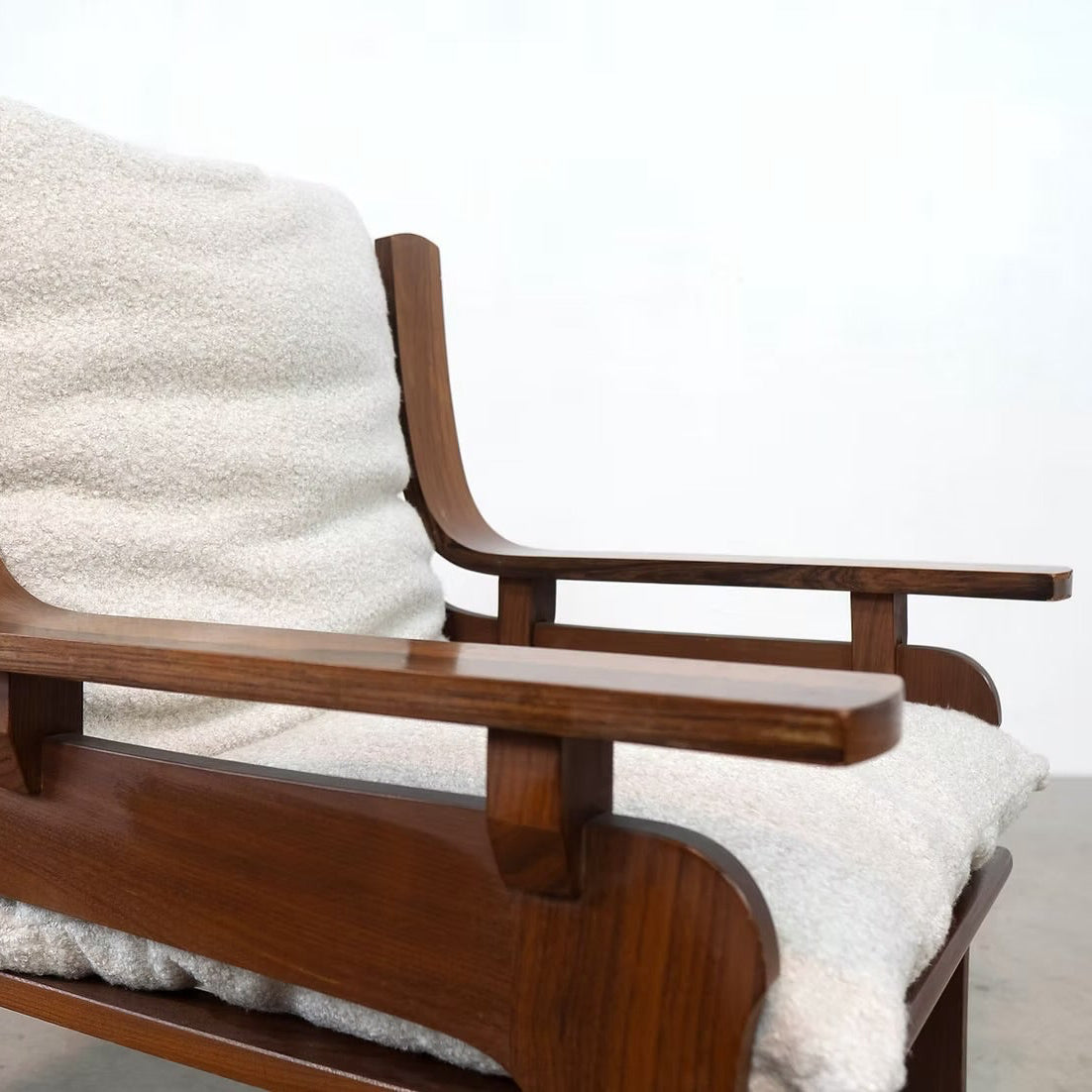 A mid-century modern wooden armchair with a curved walnut-look frame, upholstered in a textured off-white bouclé fabric, shown against a plain white wall and concrete floor.