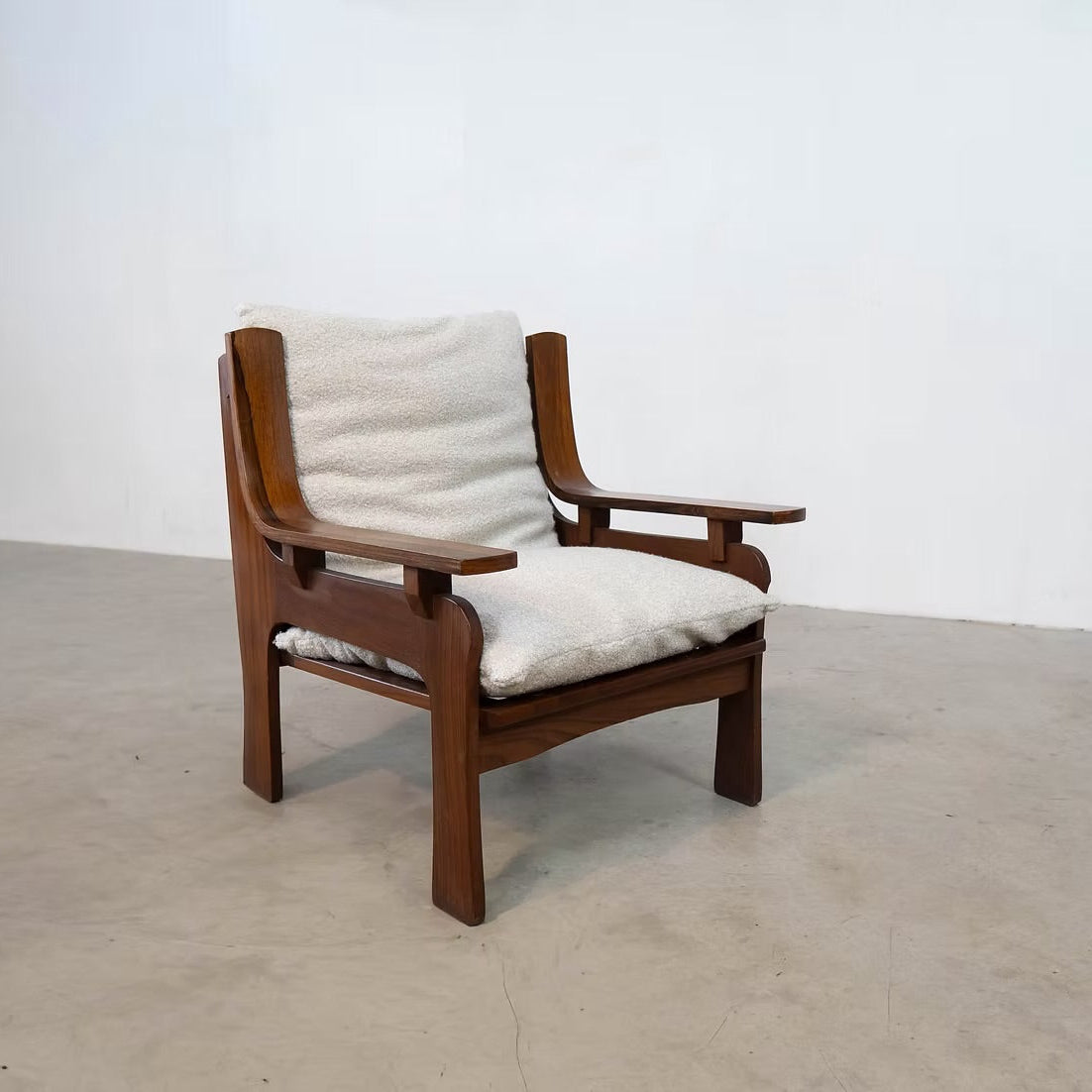 A mid-century modern wooden armchair with a warm brown wood frame and off-white textured bouclé cushions, set against a plain white wall and concrete floor.