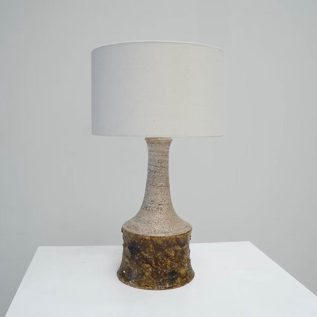 A table lamp with a textured, organic-shaped brown base and a plain white drum shade, displayed on a white pedestal against a neutral gray background.