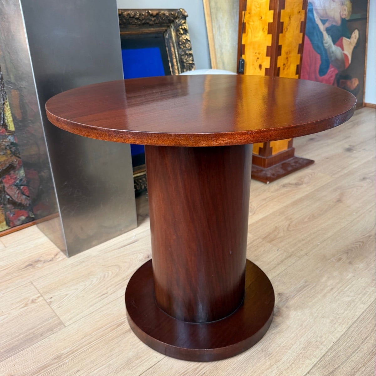 A round wooden pedestal table with a polished dark reddish-brown finish, set on a light wood floor in an indoor room with framed art and furniture in the background.