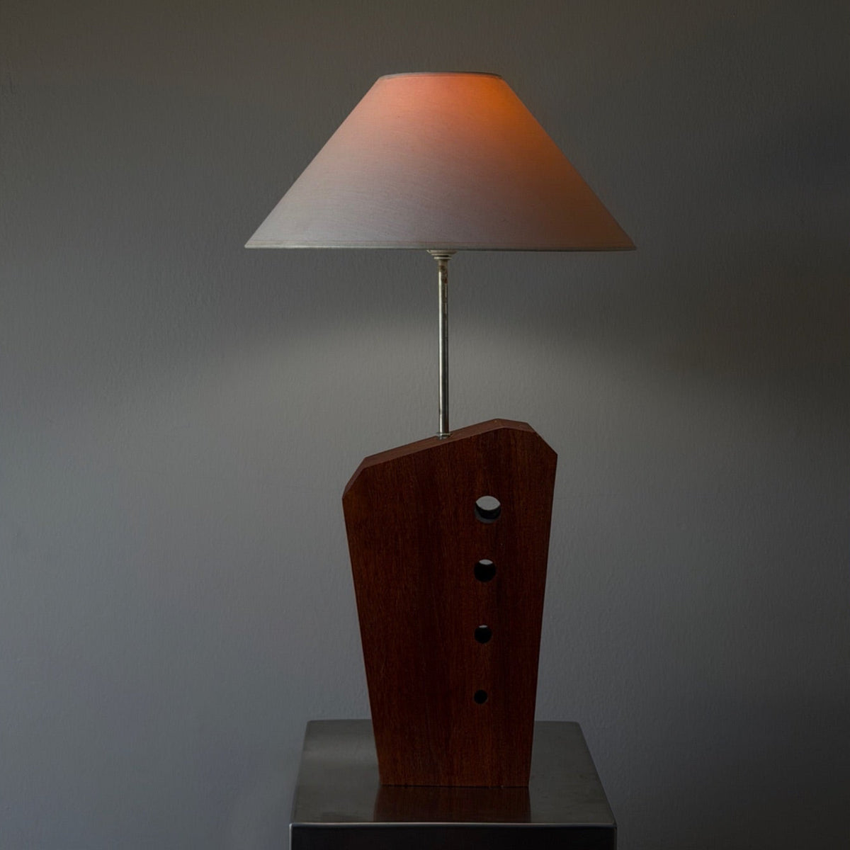 A modern table lamp with a dark brown wooden sculptural base and a tapered beige shade, shown lit against a dark gray background.