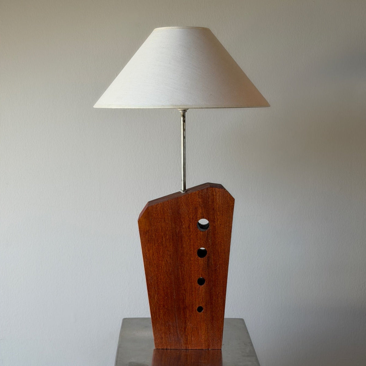A mid-century modern table lamp featuring a sculptural wooden base with a tapered white fabric shade, displayed on a metallic pedestal against a neutral background.