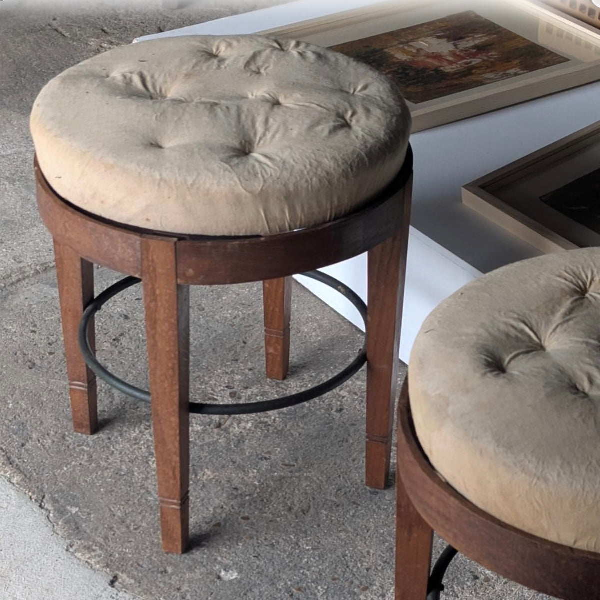 A vintage-style bar stool with a round tufted beige fabric cushion on a dark brown wooden frame, set on a concrete floor next to another matching stool.