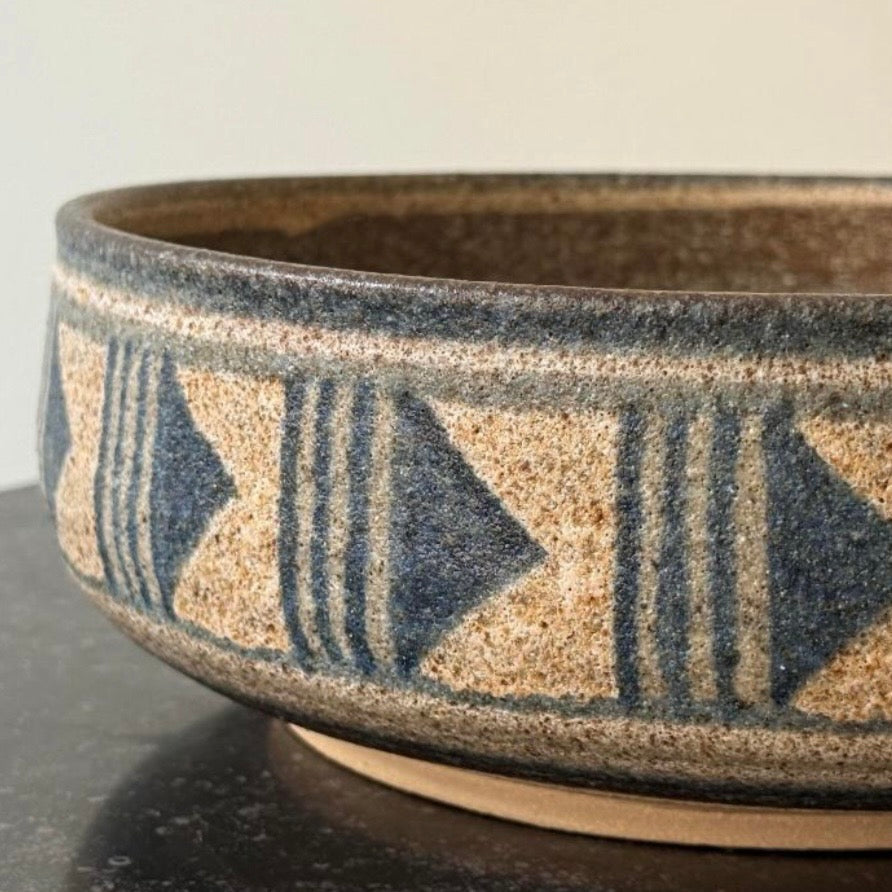 A close-up of a ceramic bowl with a textured sandy beige glaze, decorated with a dark blue geometric pattern of triangles and vertical stripes, sitting on a dark surface.