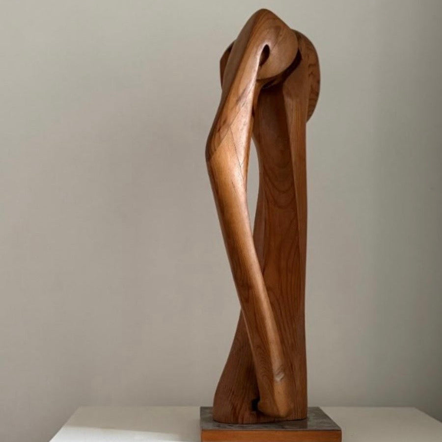 An abstract wooden sculpture with flowing, intertwined organic forms, mounted on a square base and displayed on a white pedestal against a plain light gray wall.