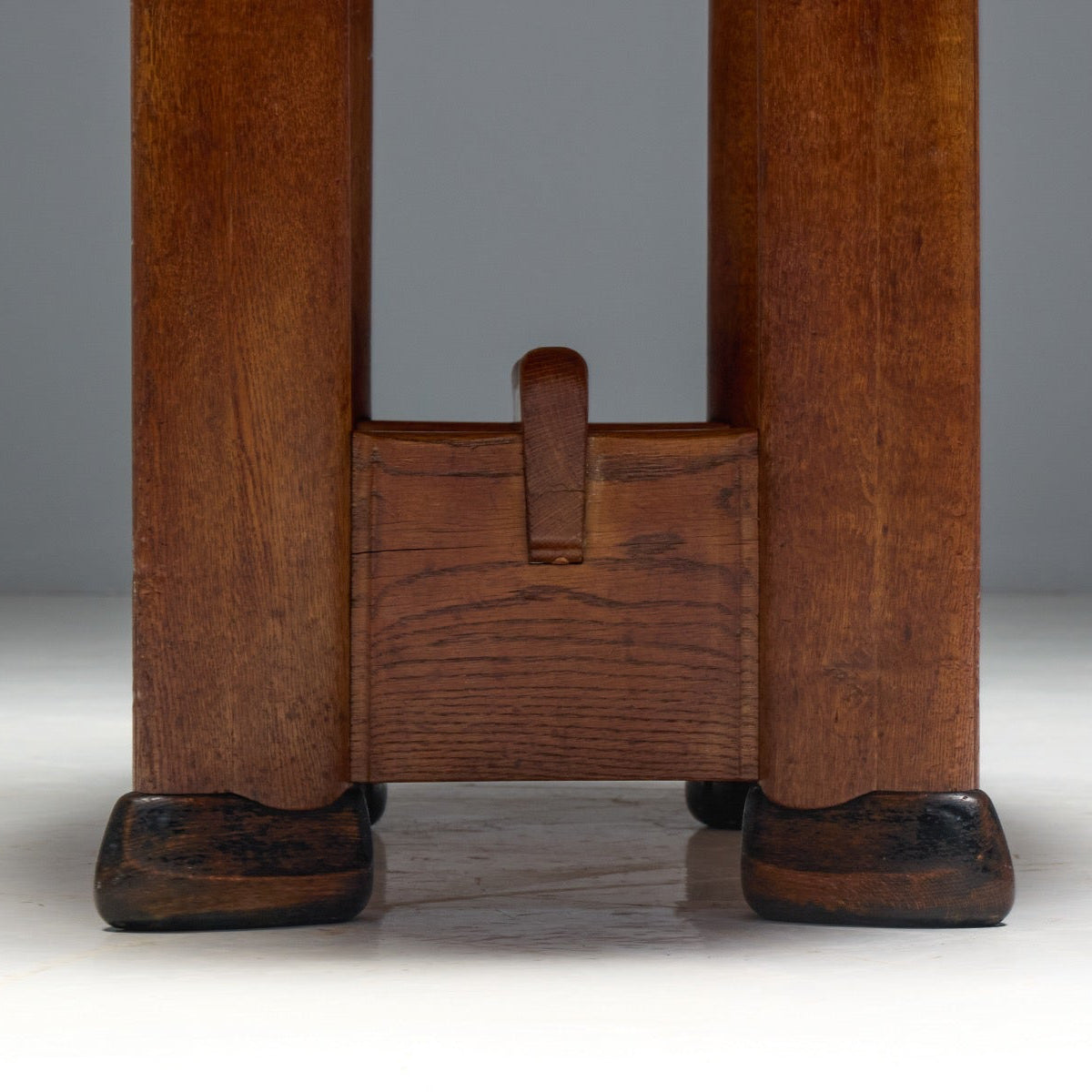 Close-up of the lower legs and feet of a dark brown wooden table or stool, showing a carved joinery detail and dark turned feet against a neutral gray studio background.