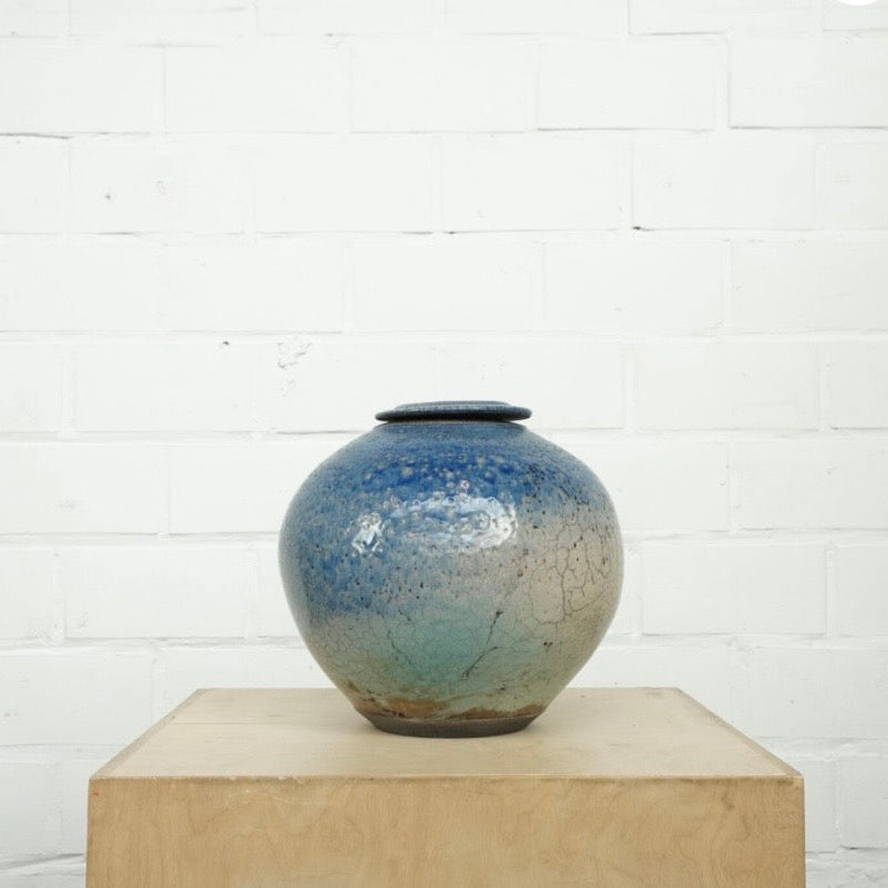 A round glazed ceramic lidded jar with a blue and green gradient glaze, displayed on a light wood block against a white brick wall.