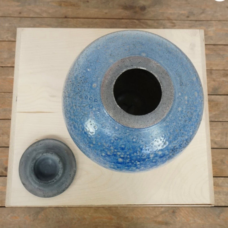 A round blue speckled ceramic vase with a matching dark gray lid, displayed on a light wood board over a rustic wooden plank floor.