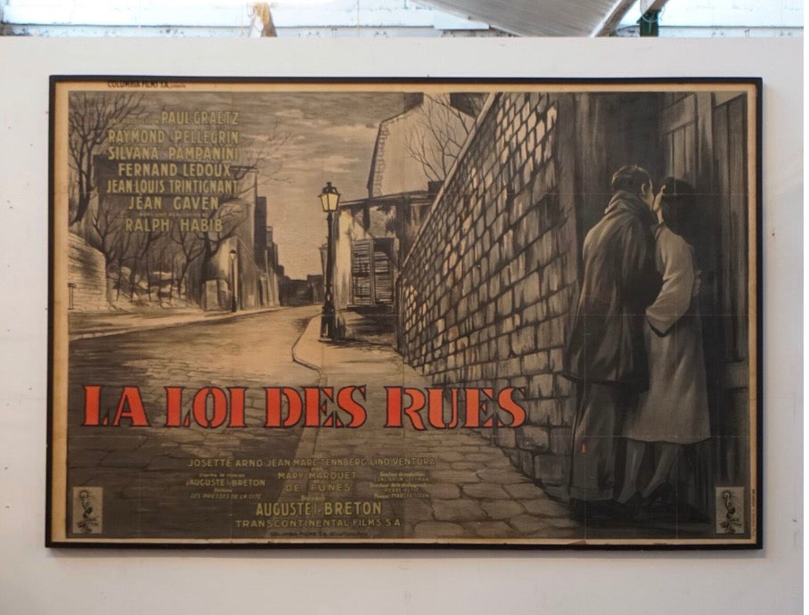 A framed vintage movie poster for the film 'La Loi des Rues' hangs on a white wall. The poster features a sepia-toned illustration of a street scene with two figures standing by a wall, and bold red title text at the bottom.