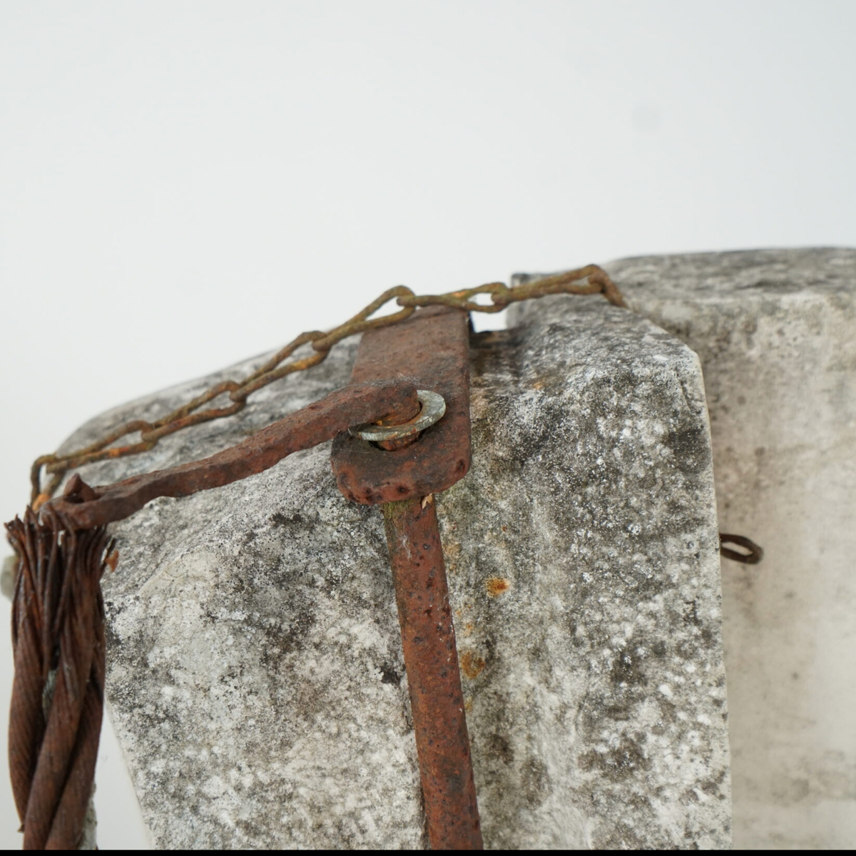 A close-up of a weathered stone or concrete object with a rusted brown strap and chain attached, shown against a plain light background.