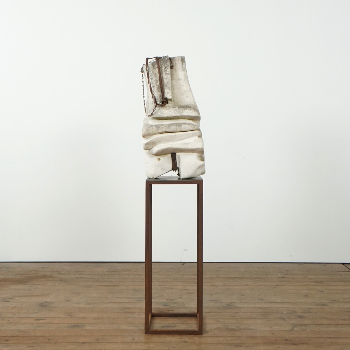 A sculptural abstract form in off-white and beige tones mounted on a tall dark metal pedestal, photographed in a gallery-like interior with a white wall and wood floor.