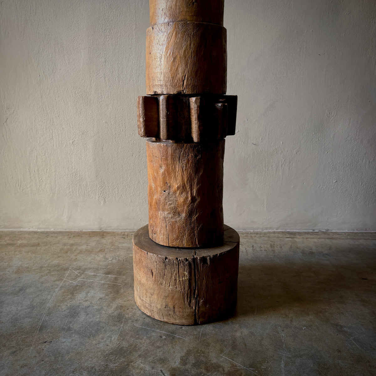 A rustic wooden sculpture or furniture piece made of stacked cylindrical and gear-like wooden sections, standing on a round wooden base against a plain textured wall and concrete floor.