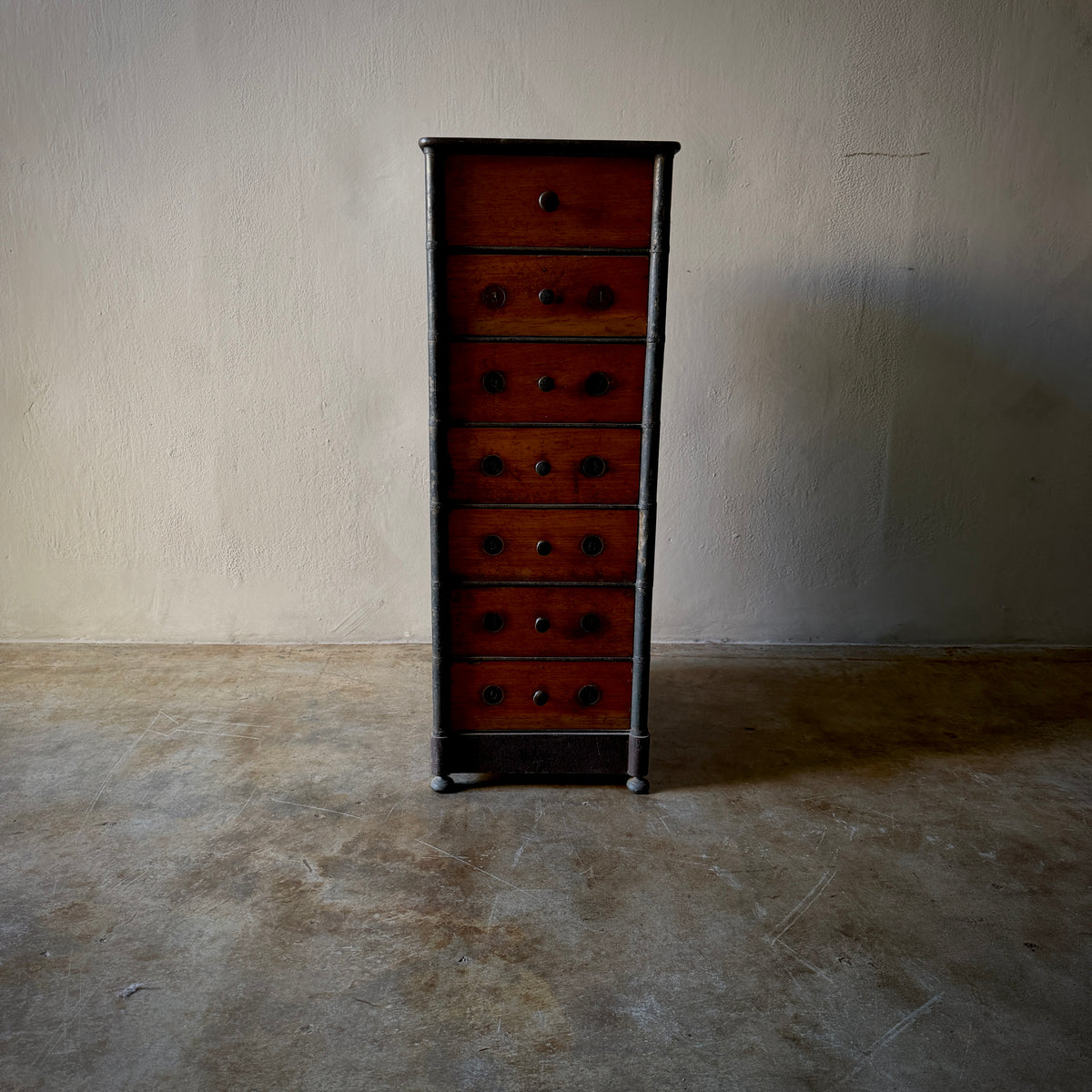 A vintage six-drawer chest of drawers with reddish-brown wood fronts and dark metal drawer pulls, set against a plain plaster wall and concrete floor.