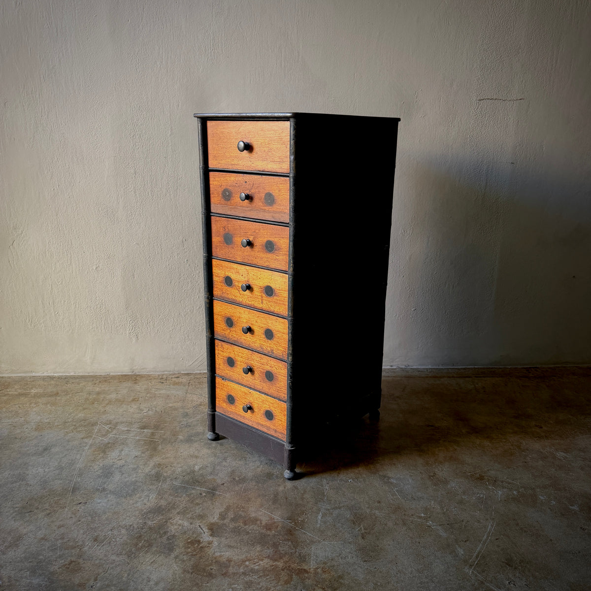 A vintage six-drawer chest of drawers with orange and brown drawers set in a black frame, standing on a concrete floor against a textured beige wall.