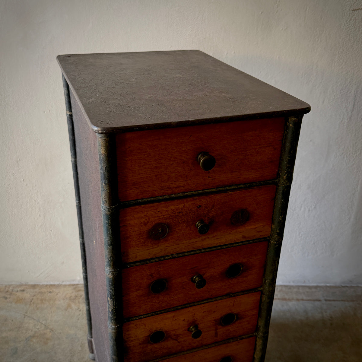 A vintage wooden chest of drawers with five drawers and dark metal knobs, set against a textured off-white wall.