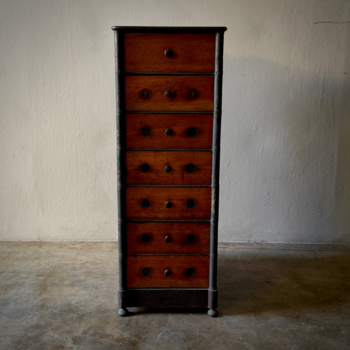 A vintage six-drawer chest of drawers with a warm reddish-brown wood finish and dark metal drawer pulls, standing on a concrete floor against a textured plaster wall.