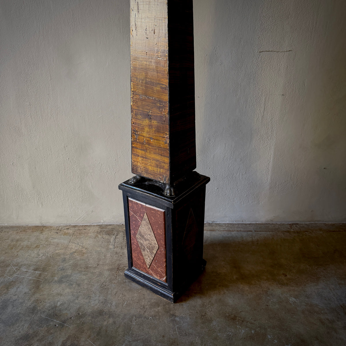 A vintage industrial column or pillar with a dark wood shaft and a black metal base featuring a reddish-brown diamond-patterned panel, set against a plain concrete floor and wall.