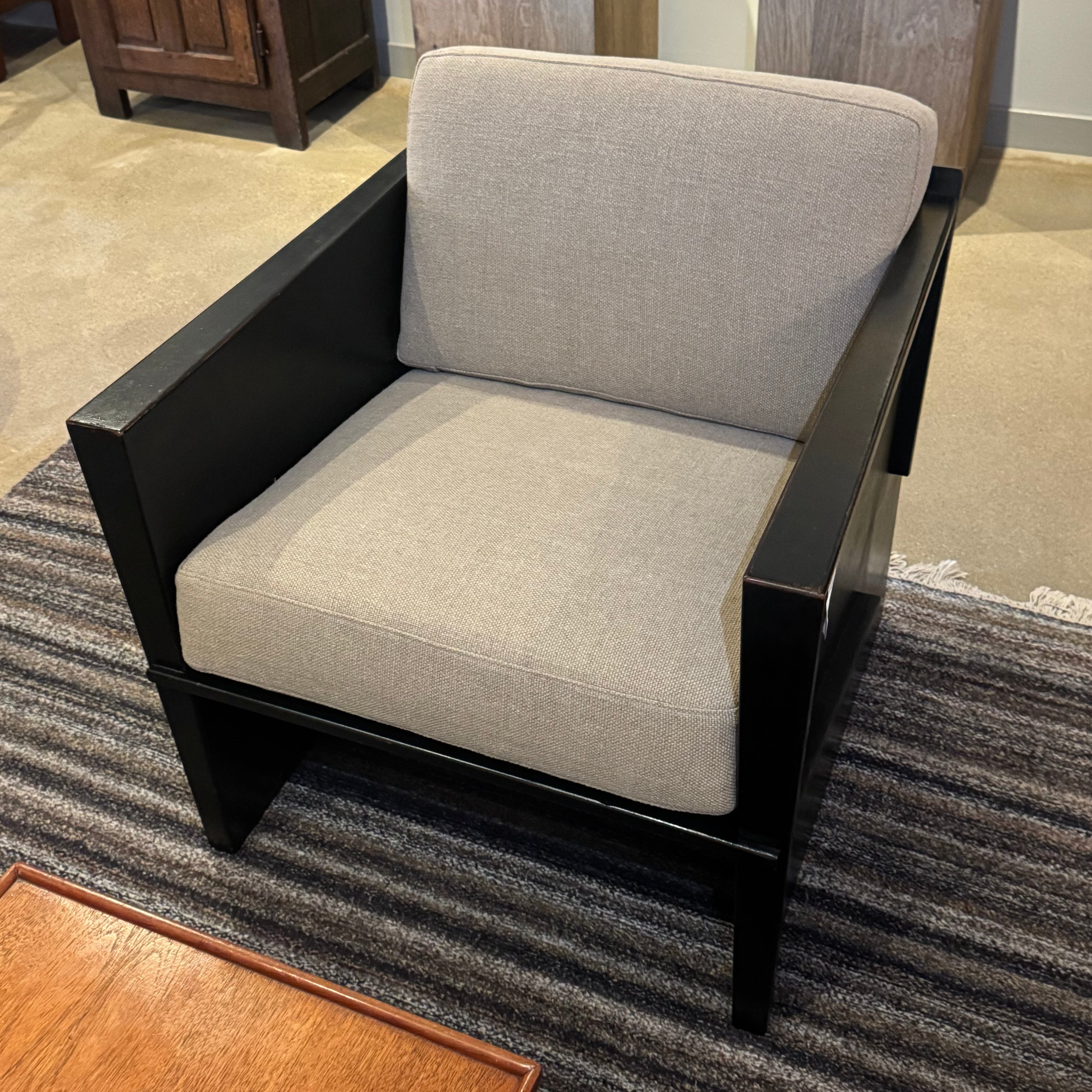 A modern accent chair with a black wooden frame and light beige textured seat and back cushions, placed on a striped gray rug in a furniture showroom.