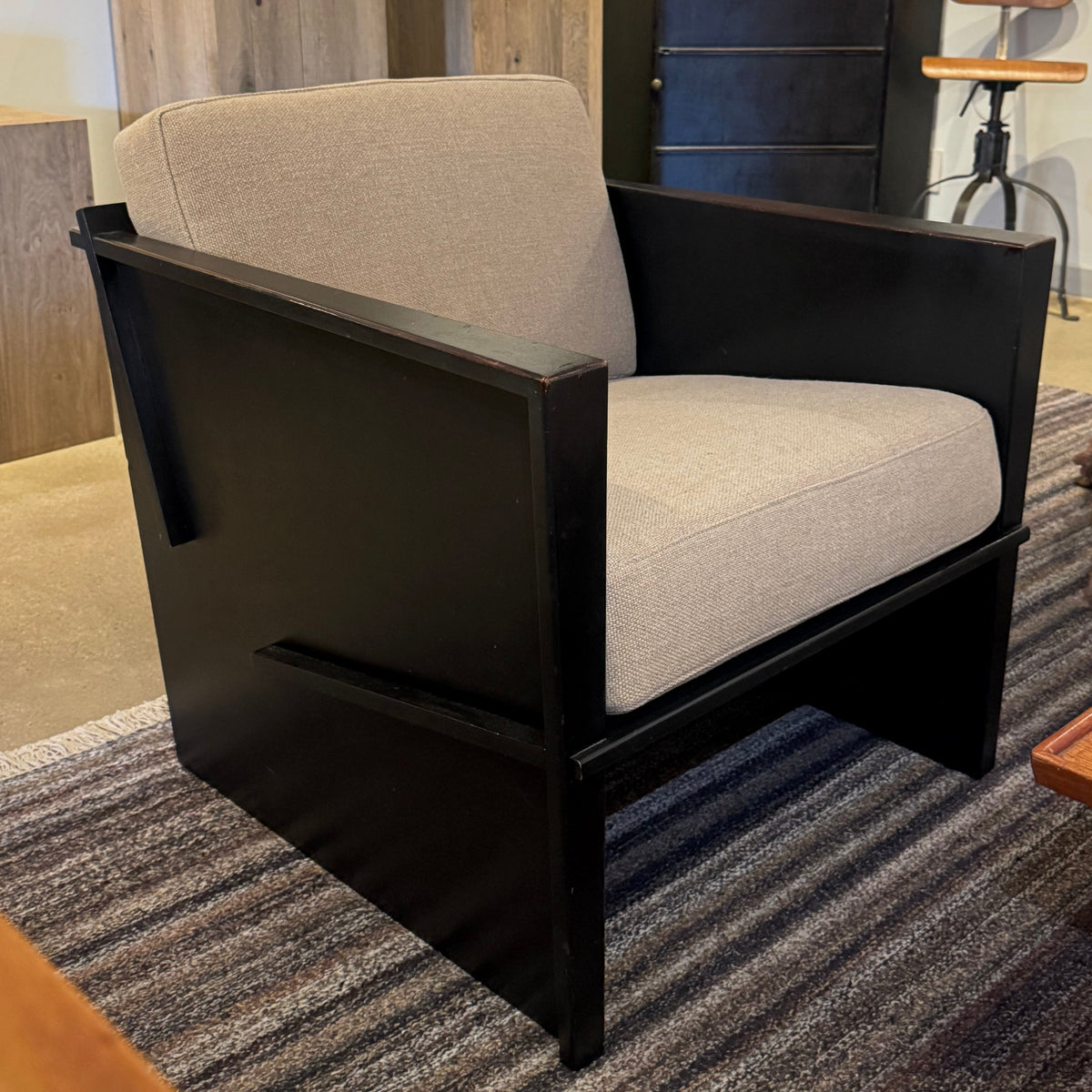 A modern accent chair with a black wooden frame and light beige textured upholstery, positioned on a striped gray rug in an indoor room setting.