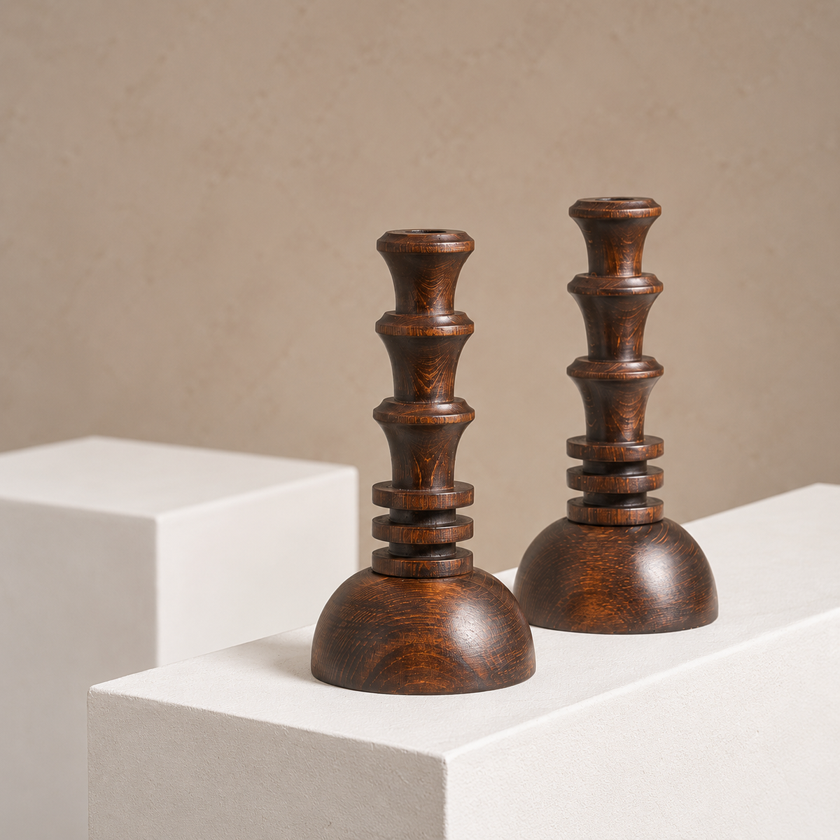 Two dark brown wooden candlesticks with a carved, tiered design and rounded bases, displayed on white pedestals against a neutral beige background.
