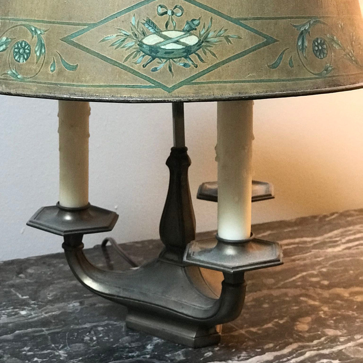 Close-up of a vintage-style table lamp with a patterned beige and teal-green shade, dual candle-style holders, and a dark metal base on a marble surface.