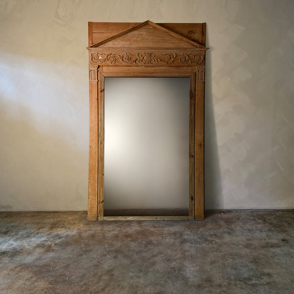 Wooden mirror frame with decorative top against a plain wall.
