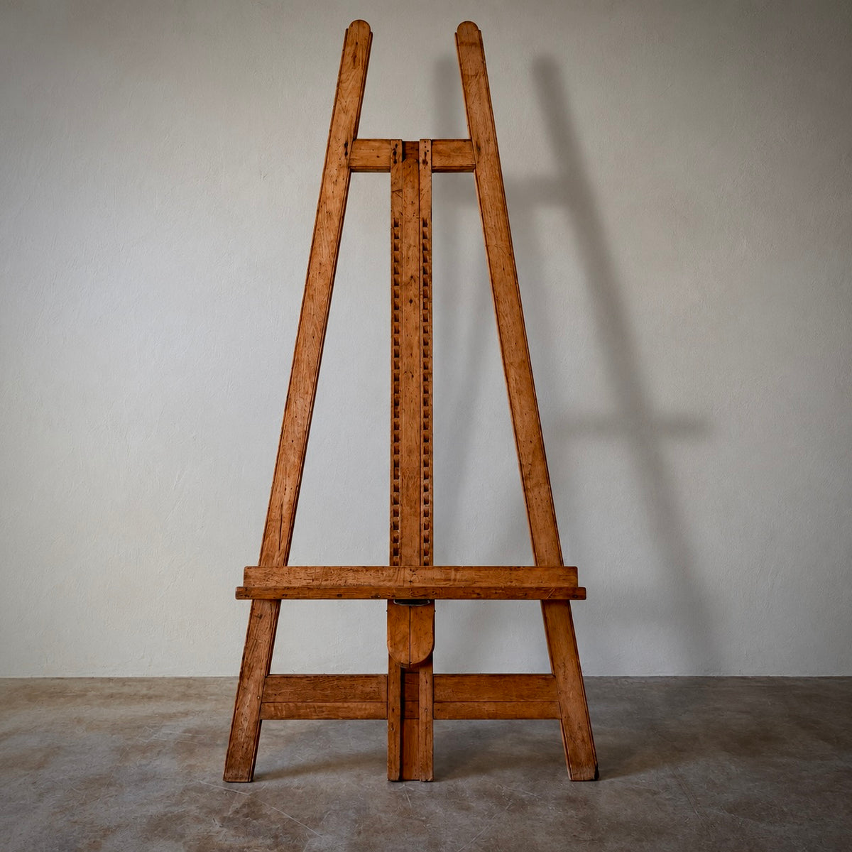 A vintage wooden artist easel made of warm brown wood, photographed from the front against a neutral gray wall and concrete floor.