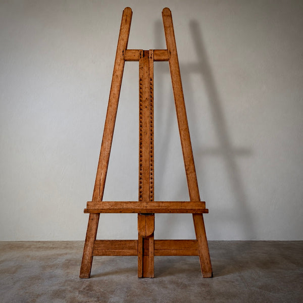 A vintage wooden artist easel made of warm brown wood, photographed from the front against a neutral gray wall and concrete floor.