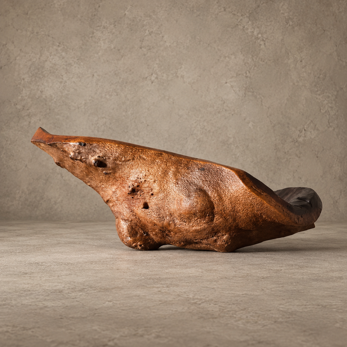 A rustic, organic-shaped brown sculpture or decorative object with a textured, weathered surface, displayed on a concrete surface against a textured gray wall.