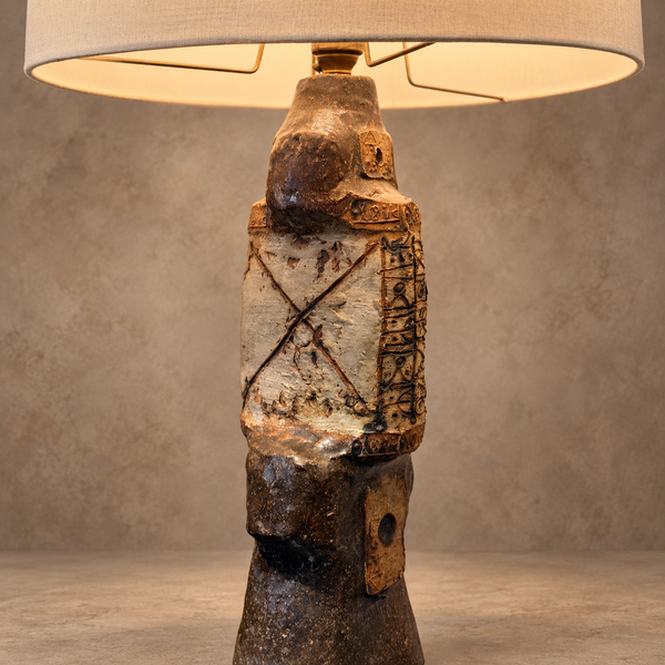 A sculptural table lamp with a textured ceramic warrior figure base and a beige drum-style shade, shown lit against a neutral textured studio background.
