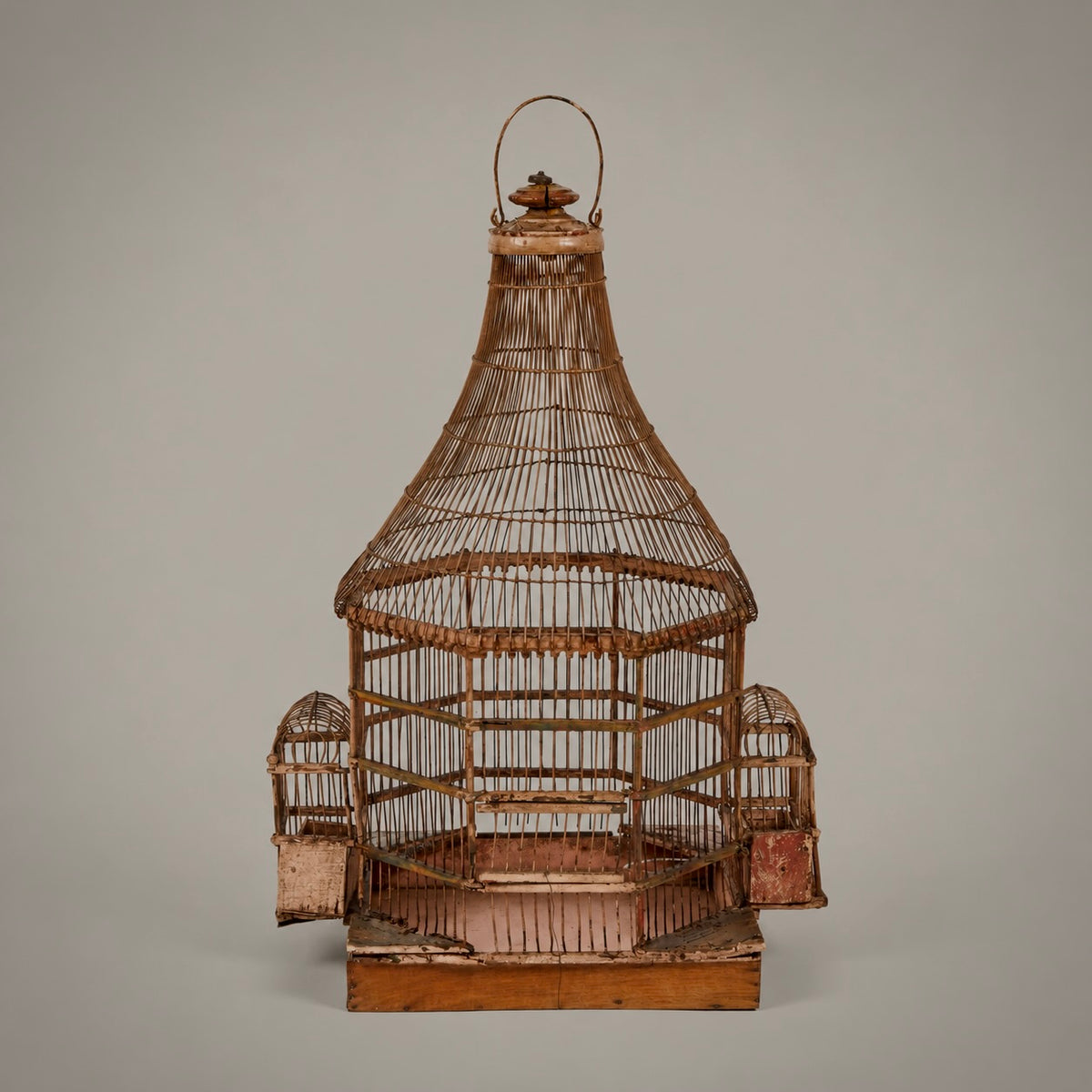 An antique wooden birdcage with a conical roof, side perches, and a hanging loop, shown against a solid gray background.