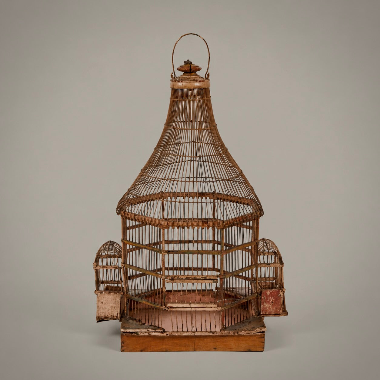An antique wooden birdcage with a conical roof, side perches, and a hanging loop, shown against a solid gray background.