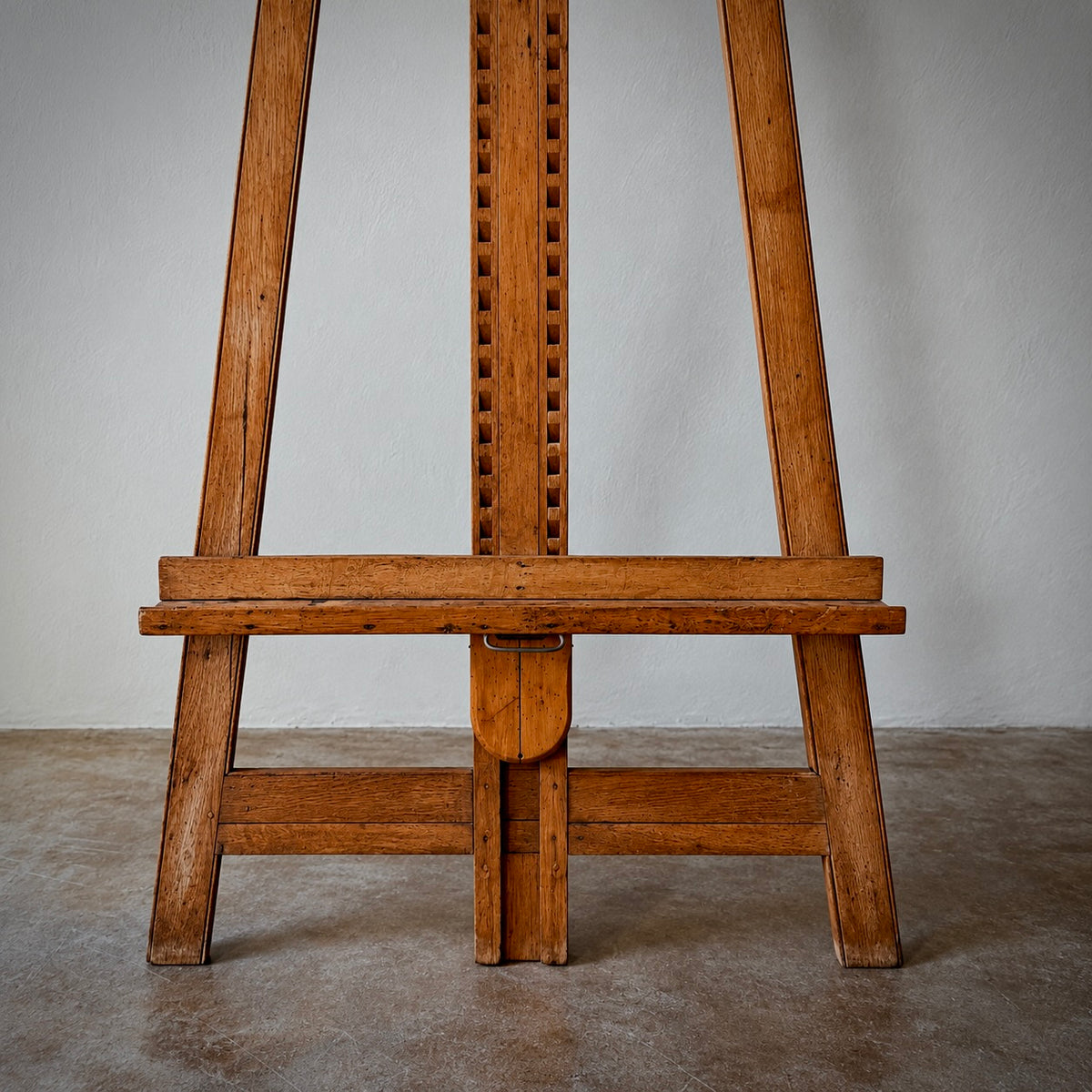 A vintage wooden artist easel made of warm brown wood, photographed from the front against a neutral gray wall and concrete floor.