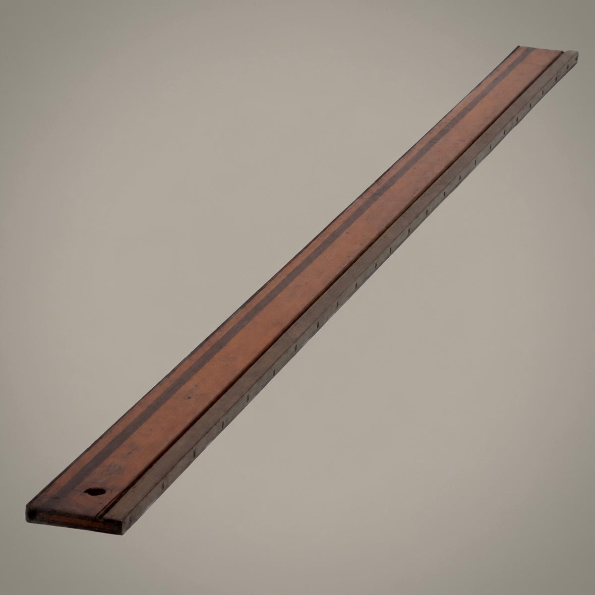 A long, narrow wooden ruler or measuring board shown diagonally on a plain warm gray background.