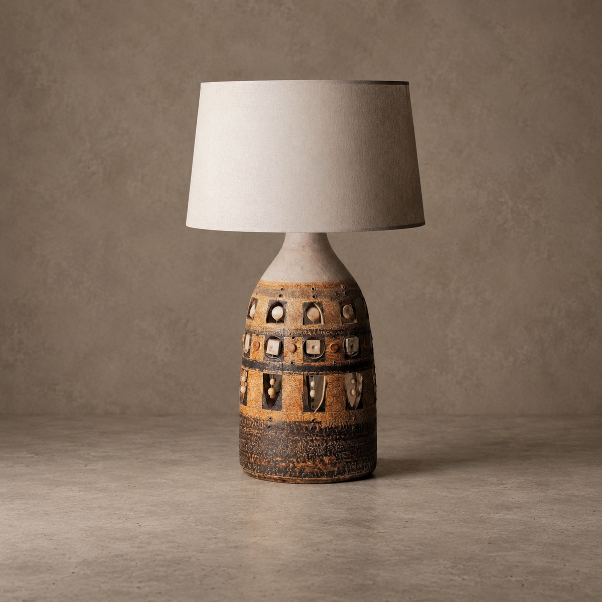 A rustic ceramic table lamp with a textured, earthy brown and beige base featuring cutout details, topped with a simple off-white fabric drum shade, set against a neutral textured studio backdrop.