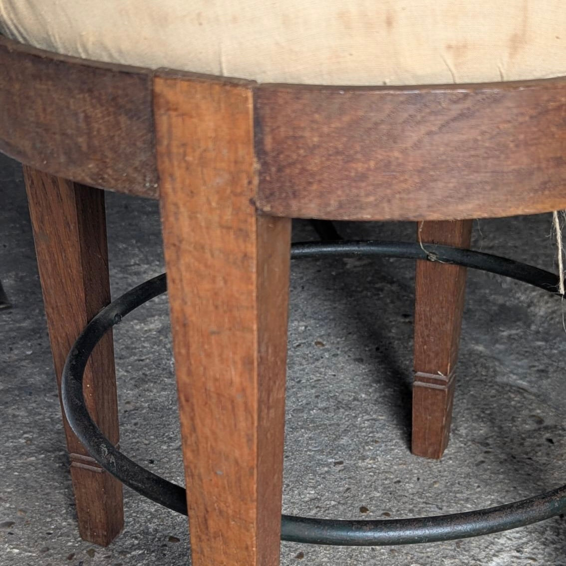 Close-up of a wooden stool with a beige upholstered seat, medium brown wooden legs, and a black metal circular footrest on a gray concrete floor.