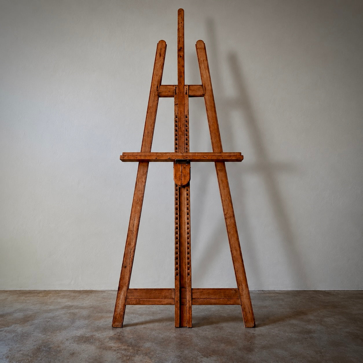A wooden easel with a warm medium-brown finish stands on a concrete floor against a neutral gray wall, casting a soft shadow to the right.