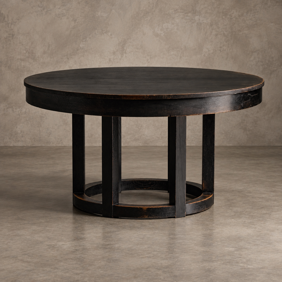 A round black wooden dining table with a distressed finish and a geometric multi-leg base, set against a textured gray concrete wall and floor.