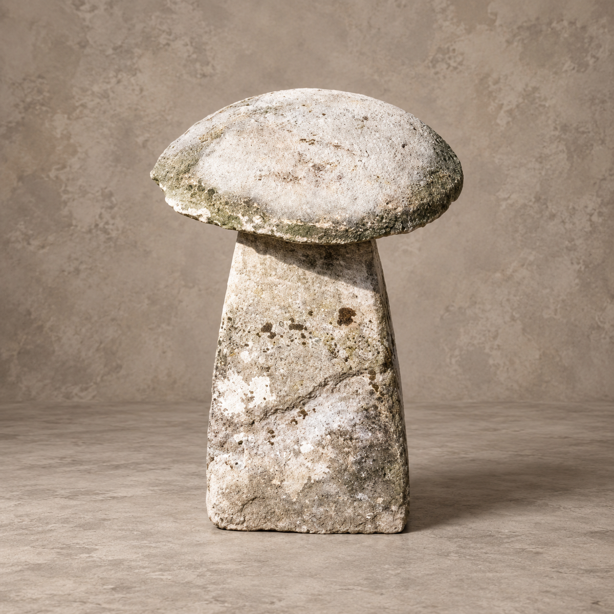A rustic stone sculpture shaped like a mushroom, featuring a textured, weathered surface with natural crevices and mossy green undertones, set against a neutral, textured studio backdrop.