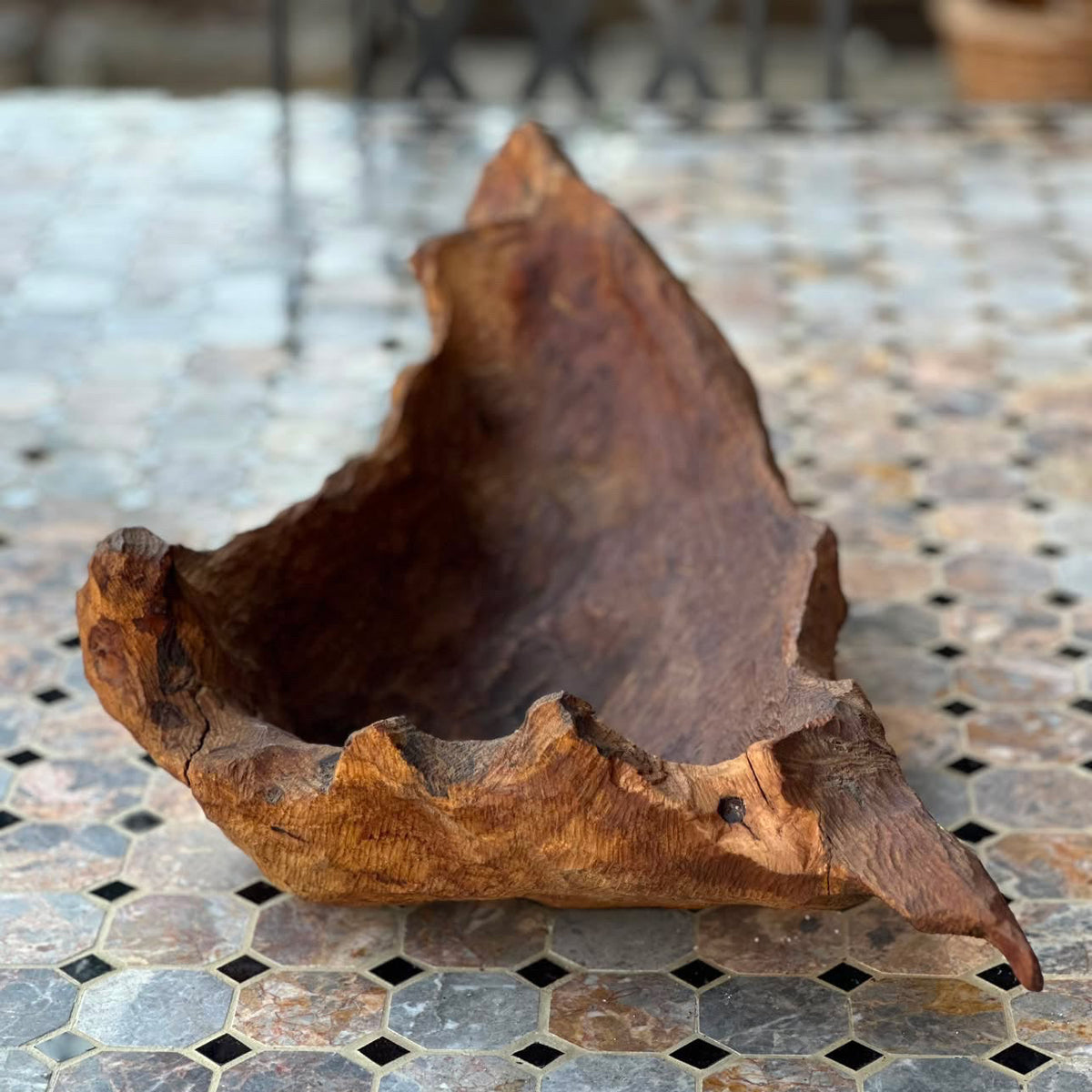 A rustic, hand-carved wooden bowl with a rough, natural bark-like rim, displayed on a mosaic tile surface.
