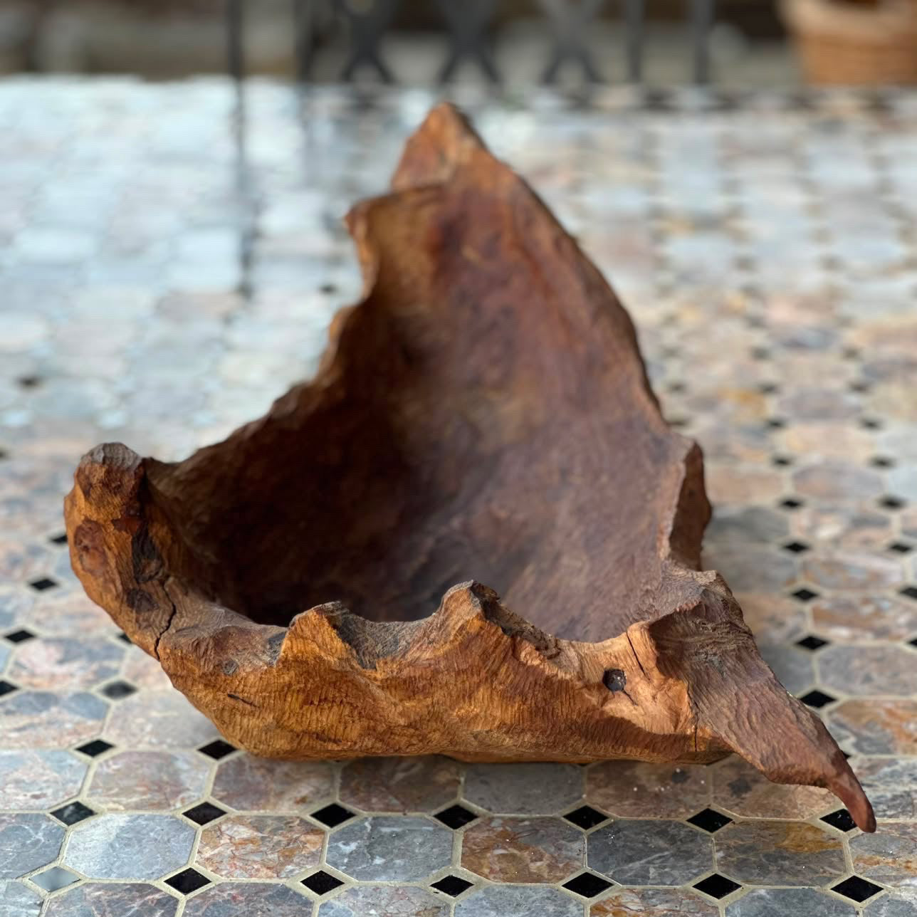 A rustic, hand-carved wooden bowl with a rough, natural bark-like rim, displayed on a mosaic tile surface.