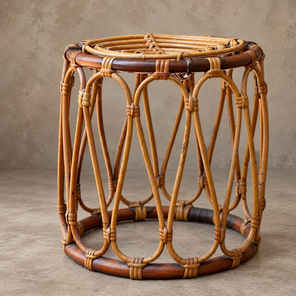 A rustic round rattan stool with a woven natural cane body and dark brown accents, set against a neutral textured concrete background.