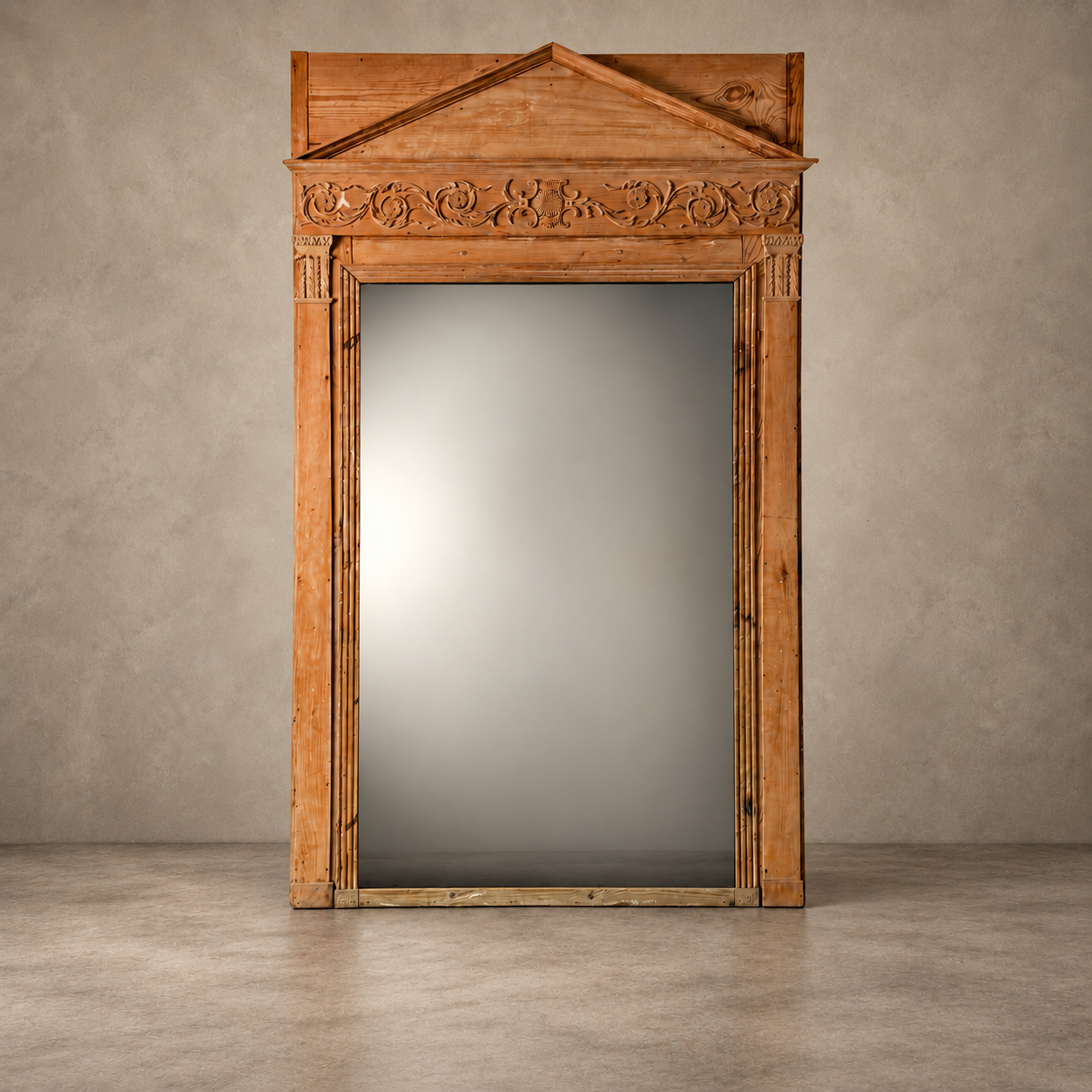 A large rectangular wall mirror with an ornate carved wooden frame and a triangular pediment top, shown standing against a textured gray wall and concrete floor.
