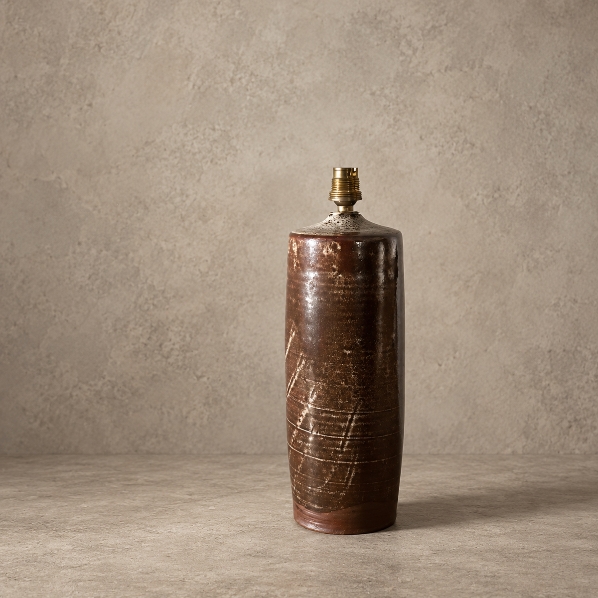 A tall, cylindrical ceramic vase with a dark brown, textured glaze and a small brass cap, set against a neutral, textured beige background.