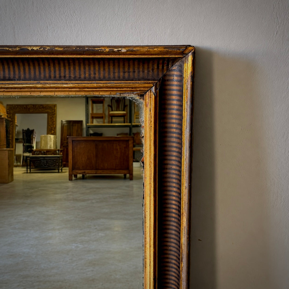 An antique-style rectangular mirror with a distressed dark brown and gold frame, leaning against a light gray wall in an antique shop.