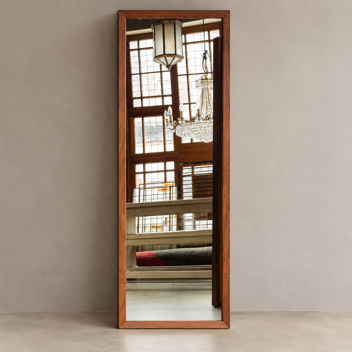 A tall, narrow wooden floor mirror with a warm medium-brown frame, leaning against a neutral gray wall in a minimalist room setting.