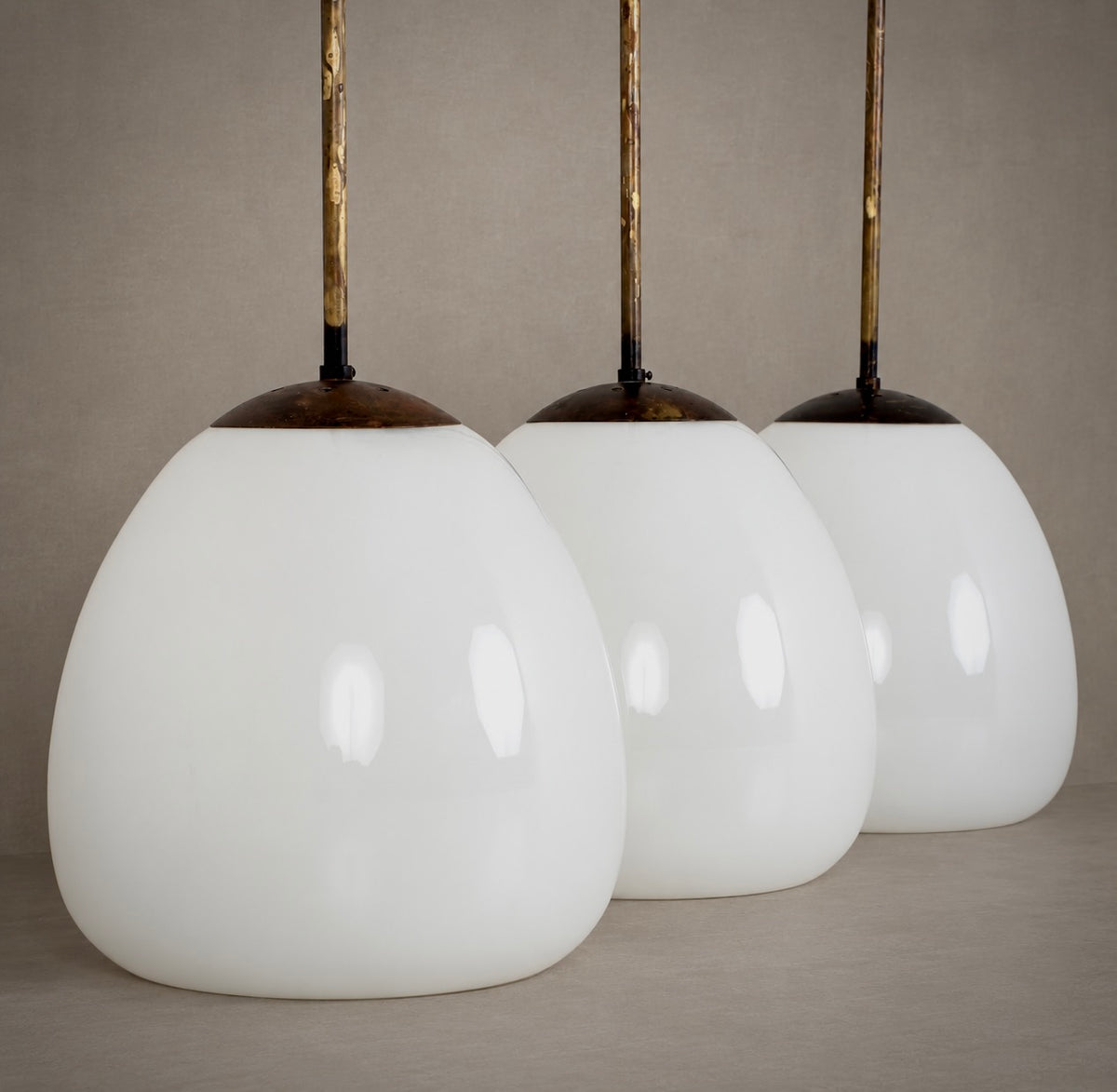 Three matching pendant light shades with large glossy white teardrop-shaped glass globes and dark metal tops, suspended by gold-toned rods against a neutral beige background.