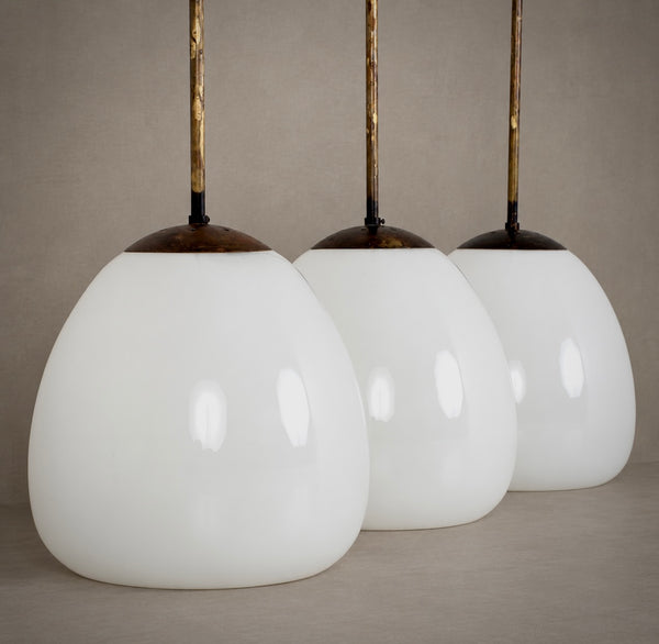Three matching pendant light shades with large glossy white teardrop-shaped glass globes and dark metal tops, suspended by gold-toned rods against a neutral beige background.